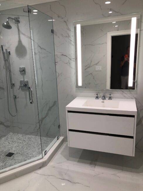 Modern bathroom featuring a glass shower enclosure with marble walls, a sleek white vanity with dual sinks, and contemporary lighting, showcasing high-quality materials ideal for renovation.